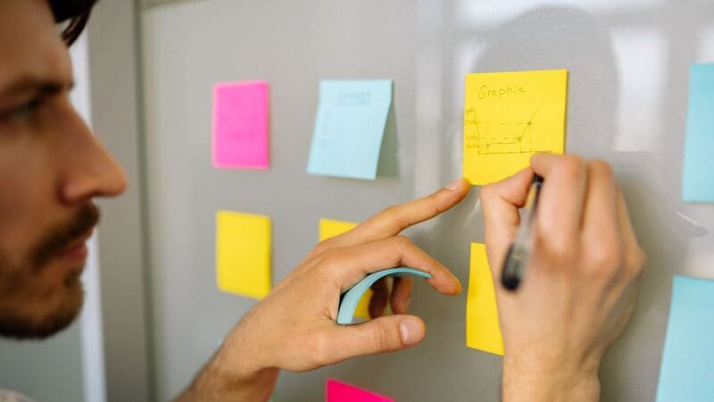 Sistema de Gestão de Projetos Rapaz escrevendo uma tarefa ou um recado em um bilhete colado na parede do escritório.